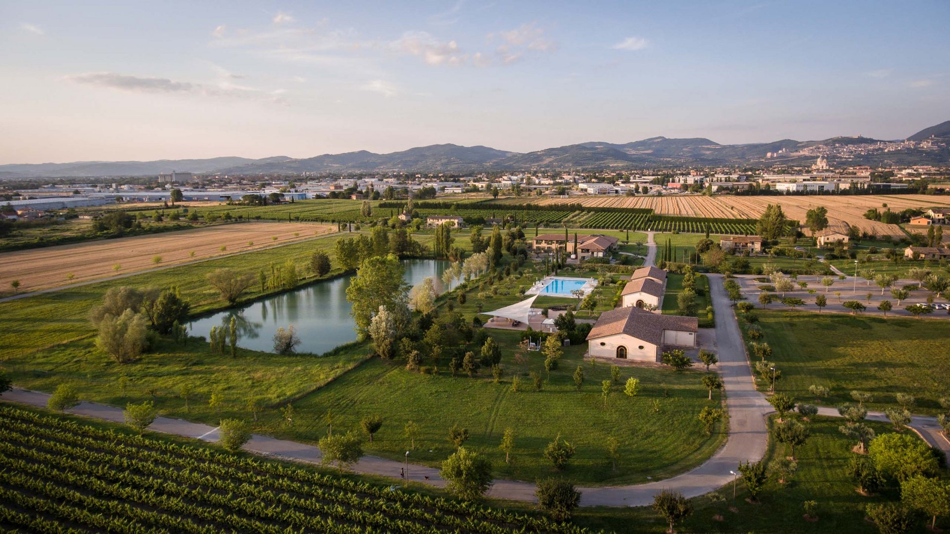 Un albergo 4 stelle e un country resort ad Assisi e i suoi proprietari Veduta aerea di un agriturismo con piscina, laghetto e campi coltivati