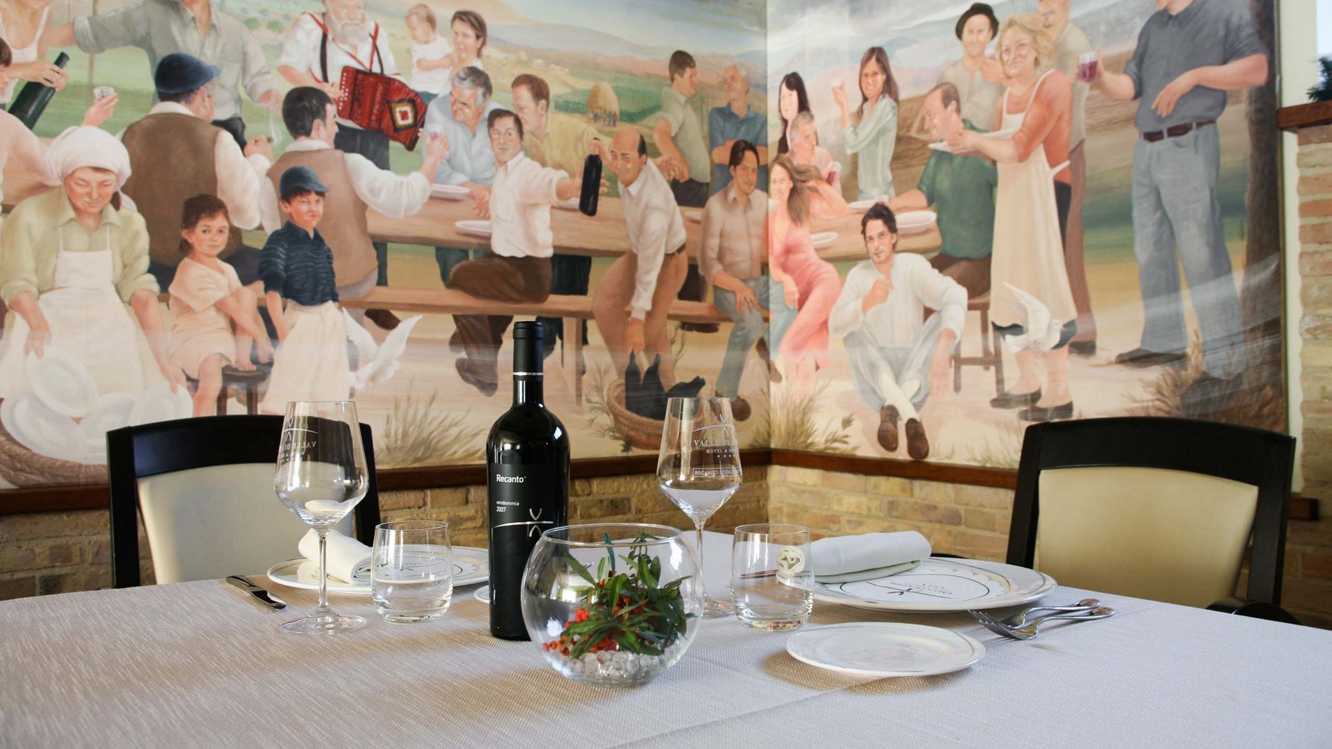 Hotel con ristorante vicino ad Assisi: benvenuti al Recanto Tavolo apparecchiato con vino davanti a murales di persone in festa