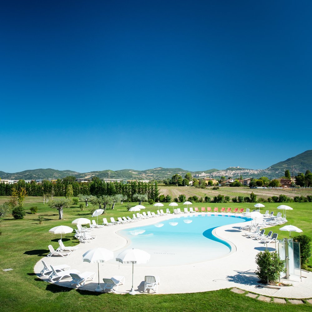 Un hotel accessibile, in Umbria, con una marcia in più Piscina a forma di fagiolo con sedie a sdraio bianche e ombrelloni in un prato verde
