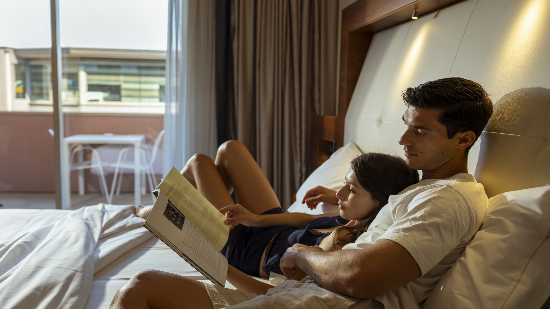 Hotel a 10 minuti di macchina da Assisi e la Basilica di San Francesco e il suo centro storico, 3 minuti di macchina dalla Porziuncola Coppia che legge insieme a letto in una stanza d'albergo con balcone